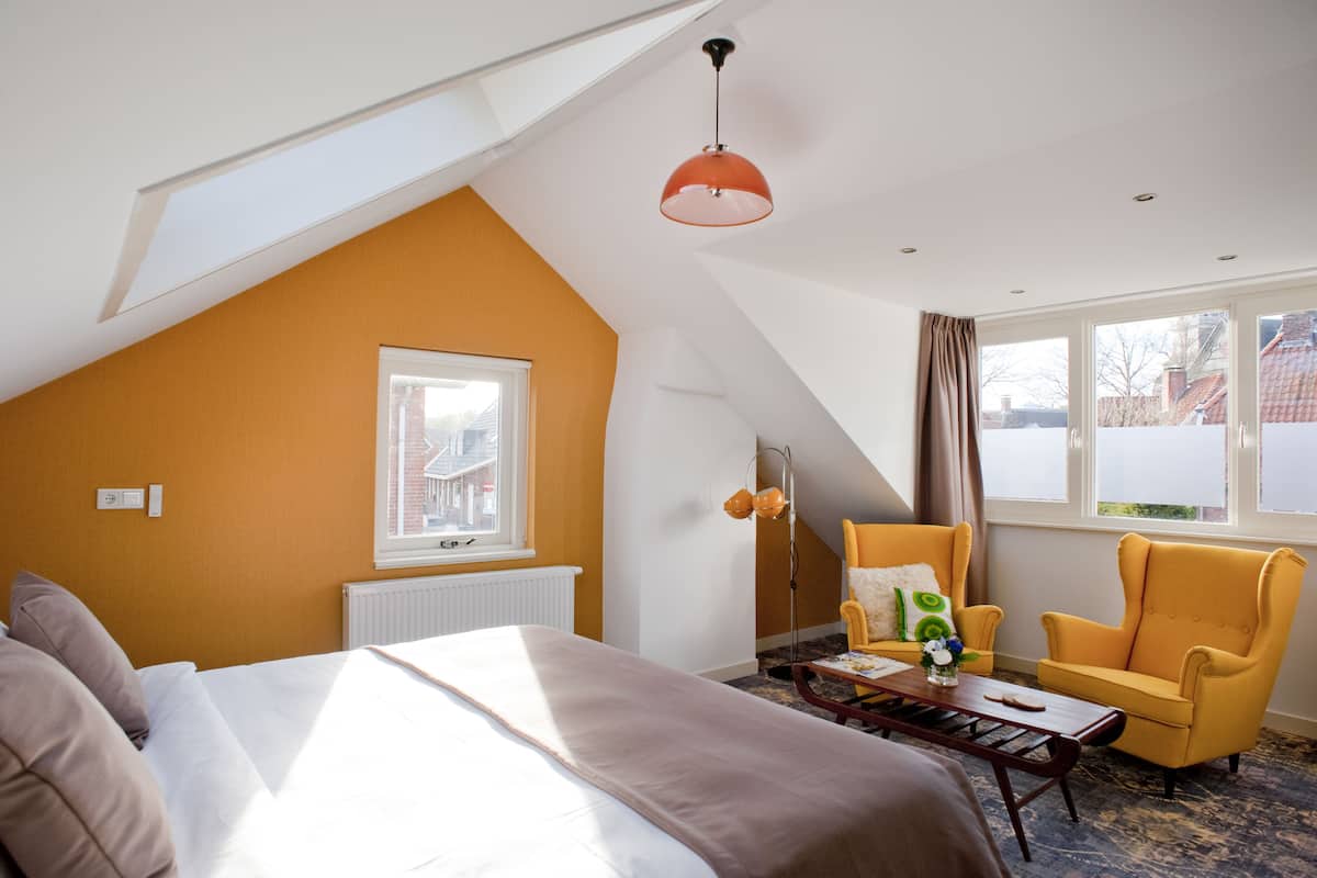Een kamer met een bed, een gele fauteuil, een tafel met een vaas en een raam met gordijnen.