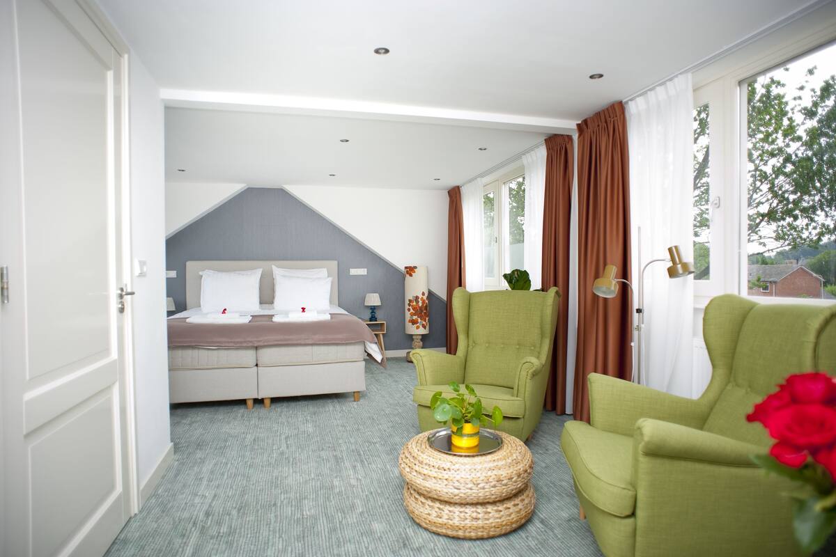 Een moderne hotelkamer met een groot bed, twee groene fauteuils, een klein rond tafeltje met een plant, en uitzicht op het landschap.