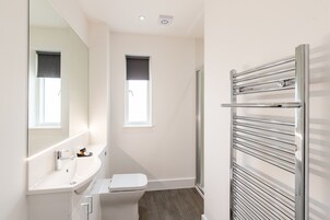 Un baño moderno con lavamanos, espejo y retrete blancos, una ducha con puerta de vidrio y un perchero para toallas.