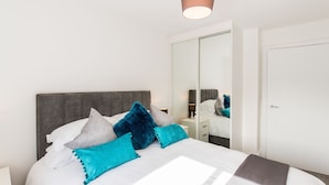 A neatly made bed with a white and grey headboard, multiple pillows, and a brown throw blanket. A small bedside table with a lamp is visible. - Abodebed - Oval View (Hemel Hempstead)