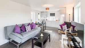 A modern living room with a grey sofa, a coffee table, and a dining area with a chair and table. - Abodebed - Oval View (Hemel Hempstead)