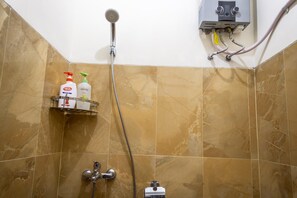 A shower area with a handheld showerhead, a wall-mounted control panel, and a shelf with toiletry bottles. - Zenrra House (Bandung)