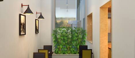 A dining area with a green artificial grass floor, wooden tables, and chairs, framed by a large window with a view of greenery.