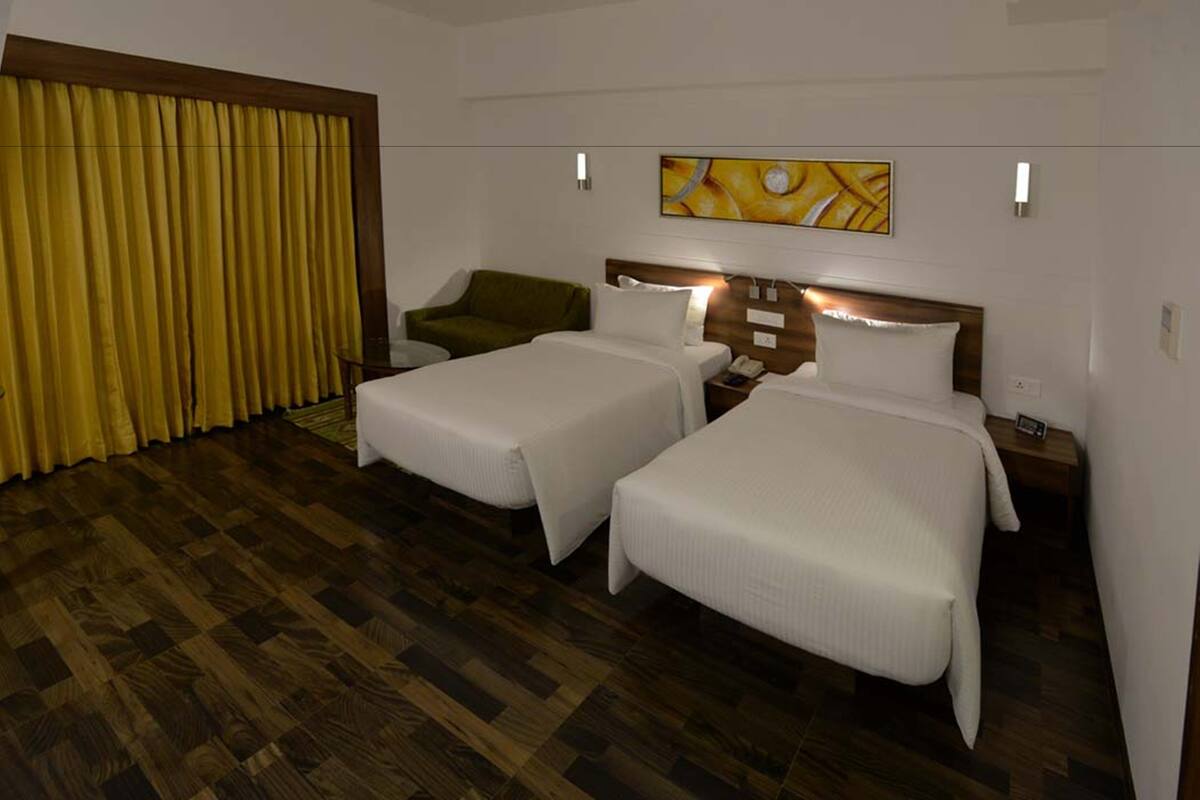 A hotel room with two single beds, a green chair, a wooden headboard, and a yellow curtain.