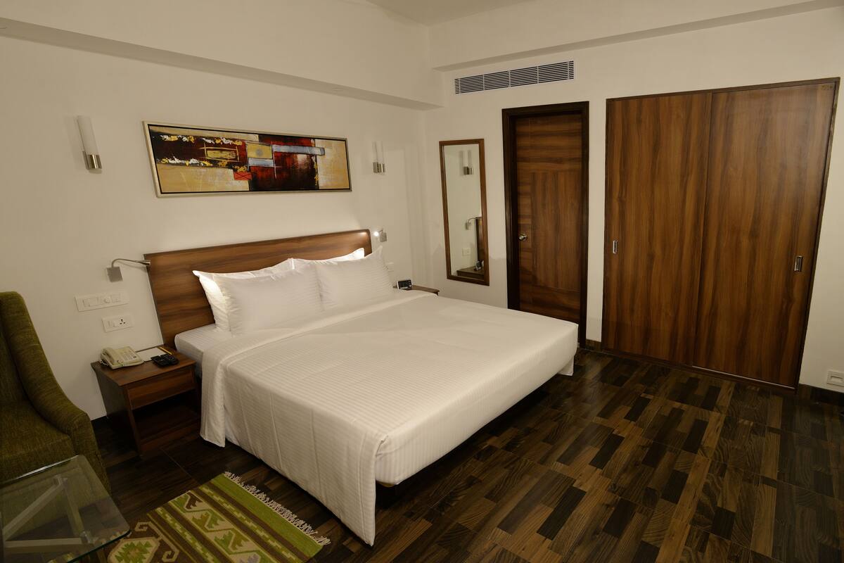 A hotel room with a large bed, a wooden wardrobe, a bedside table with a lamp, a chair, and a painting on the wall.