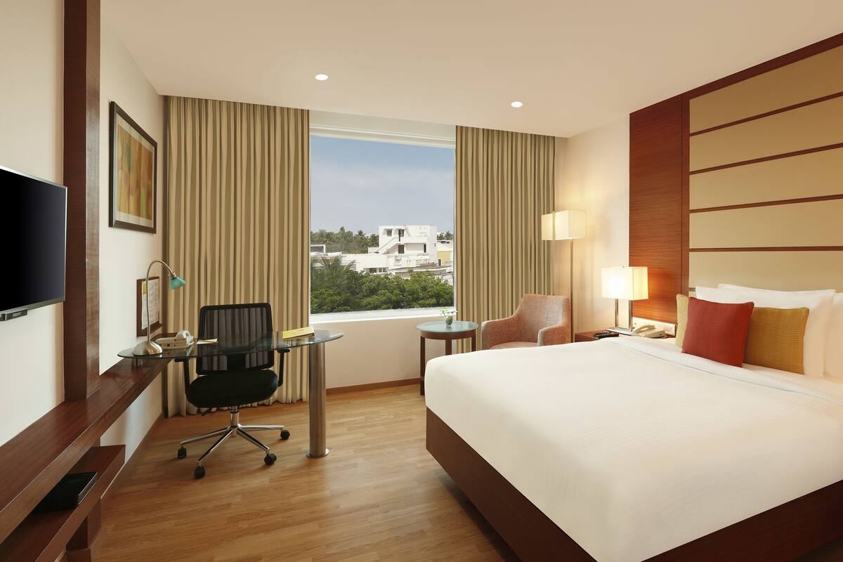 A hotel room with a large bed, a desk with a chair, a TV, a window with curtains, and a view of buildings outside.