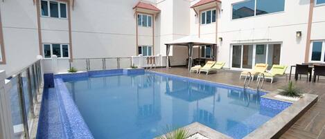 Una piscina con borde de azulejos, rodeada de sillas de descanso y un área de estar cubierta.