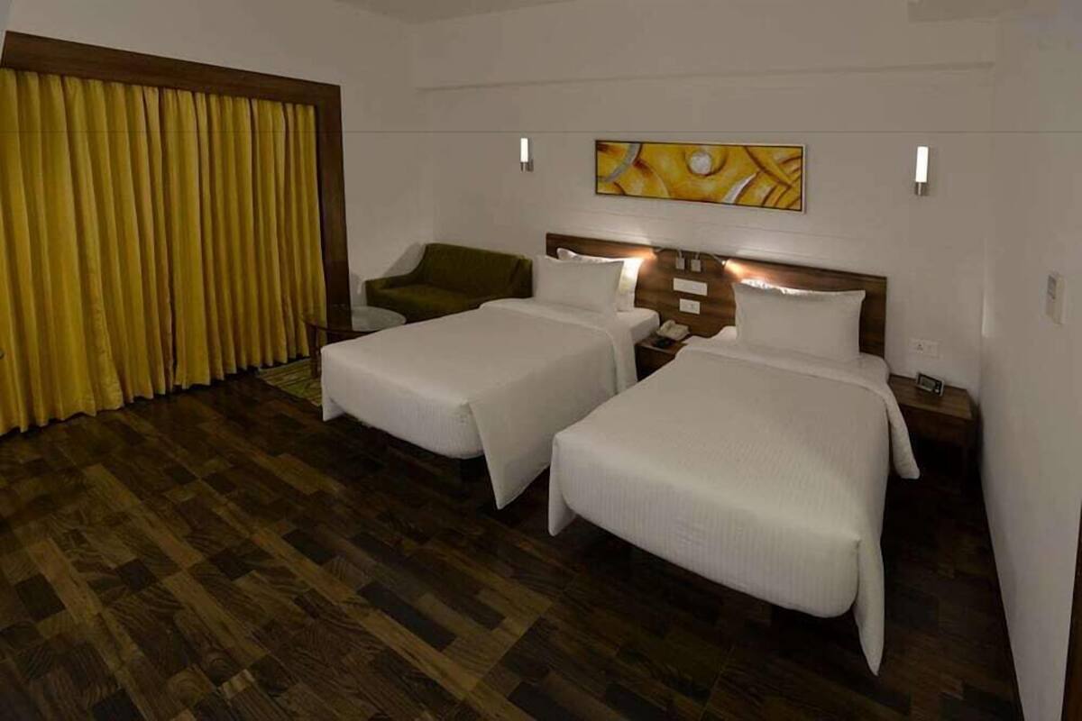 A hotel room with two beds, a green chair, a wooden headboard, and a yellow curtain.