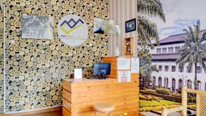 Un mostrador de recepción con detalles de madera, un logotipo y un cartel. Hay una silla y una mesita con un ventilador.