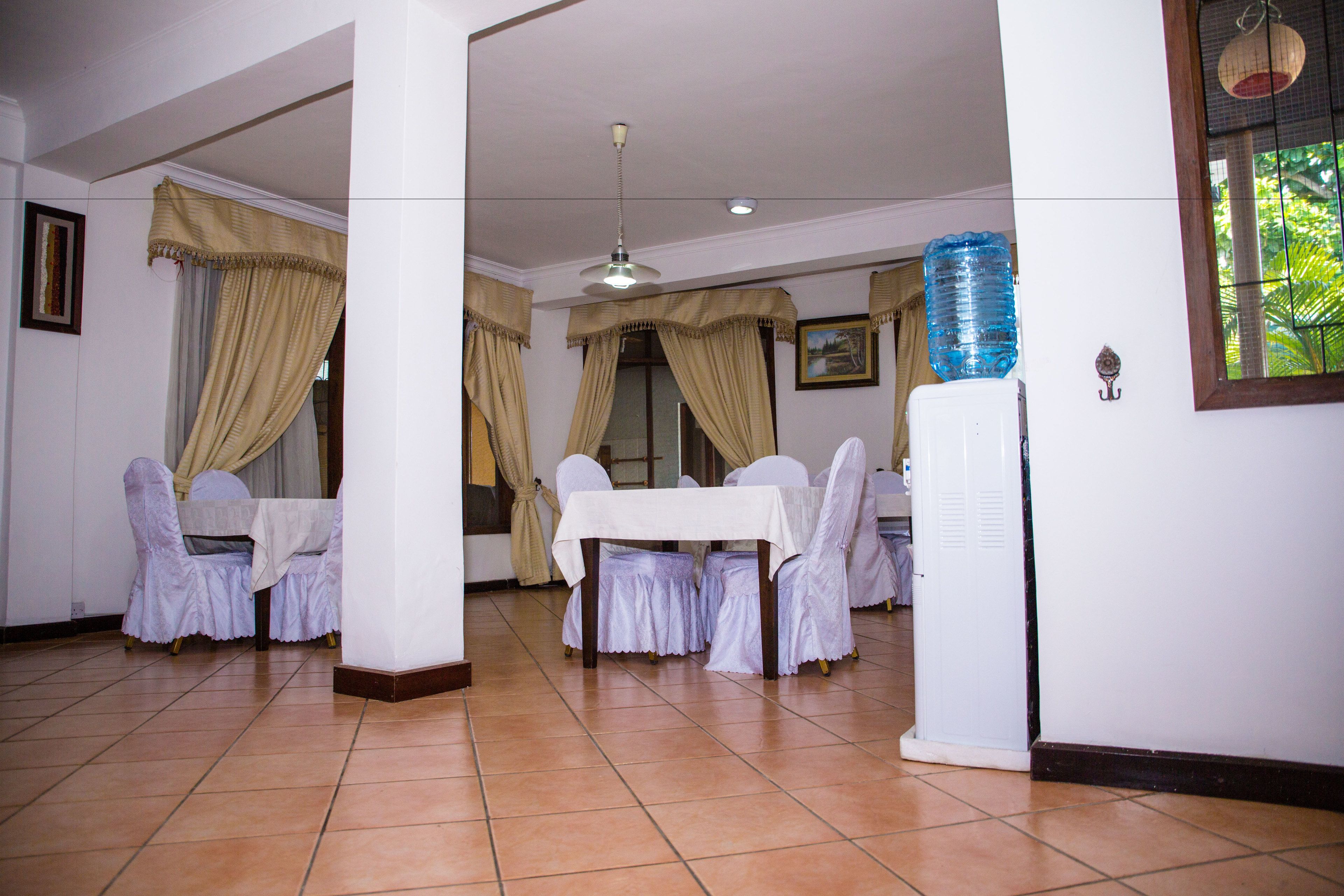 Un comedor con mesas y sillas blancas, cortinas color crema y suelo de baldosas.
