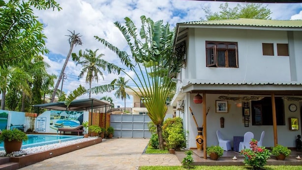 Une maison à deux étages avec une piscine, entourée de plantes tropicales et dotée d’un patio couvert.