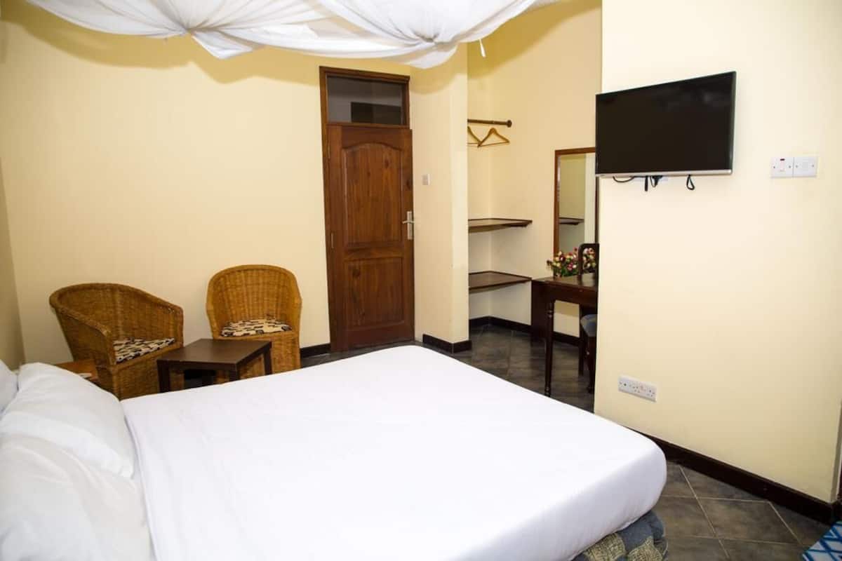 Ein Hotelzimmer mit einem Bett, zwei Korbstühlen, einer Holztür, einem an der Wand befestigten Fernseher und einem kleinen Tisch mit einer Vase.