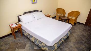 A neatly made bed with white linens, a small wooden nightstand with a phone and a magazine, and two wicker chairs with cushions.