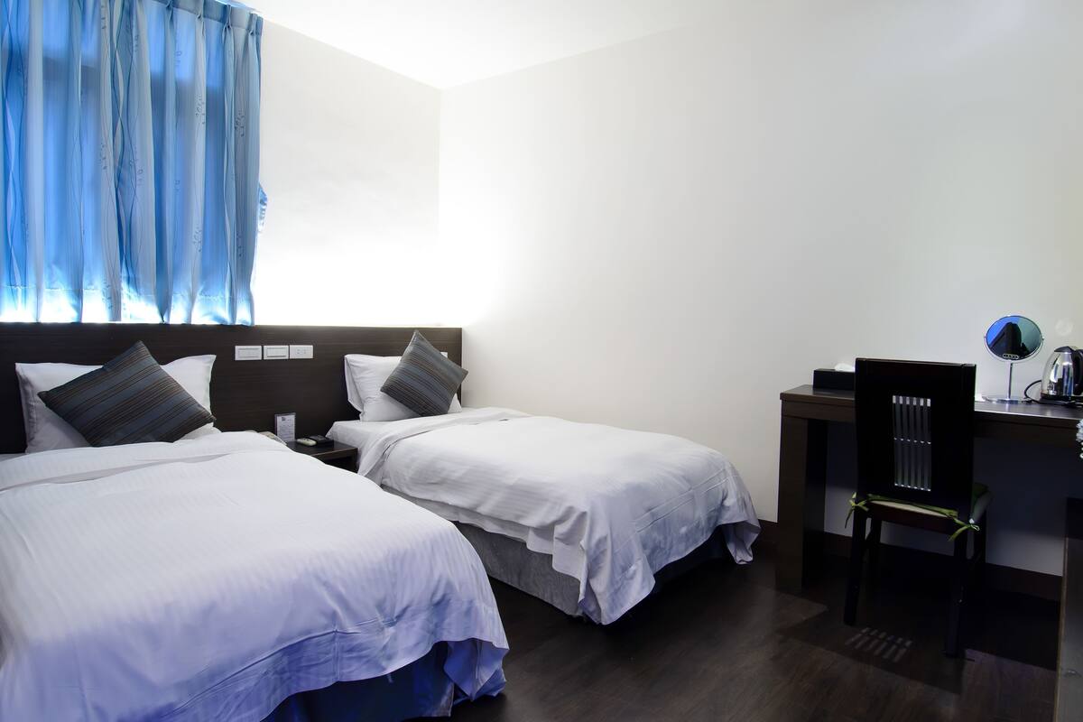 A hotel room with two single beds, a desk, a chair, and a window with blue curtains.
