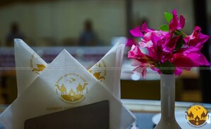 Ein Tisch mit einer weißen Serviette, dem Logo vom Desert Adventure Camp und einer Vase mit rosa Blumen.