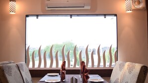 A dining area with a table set for two, a window with a view of trees, and patterned chairs. - Ashoka Residency Hotel (Bhilwara)