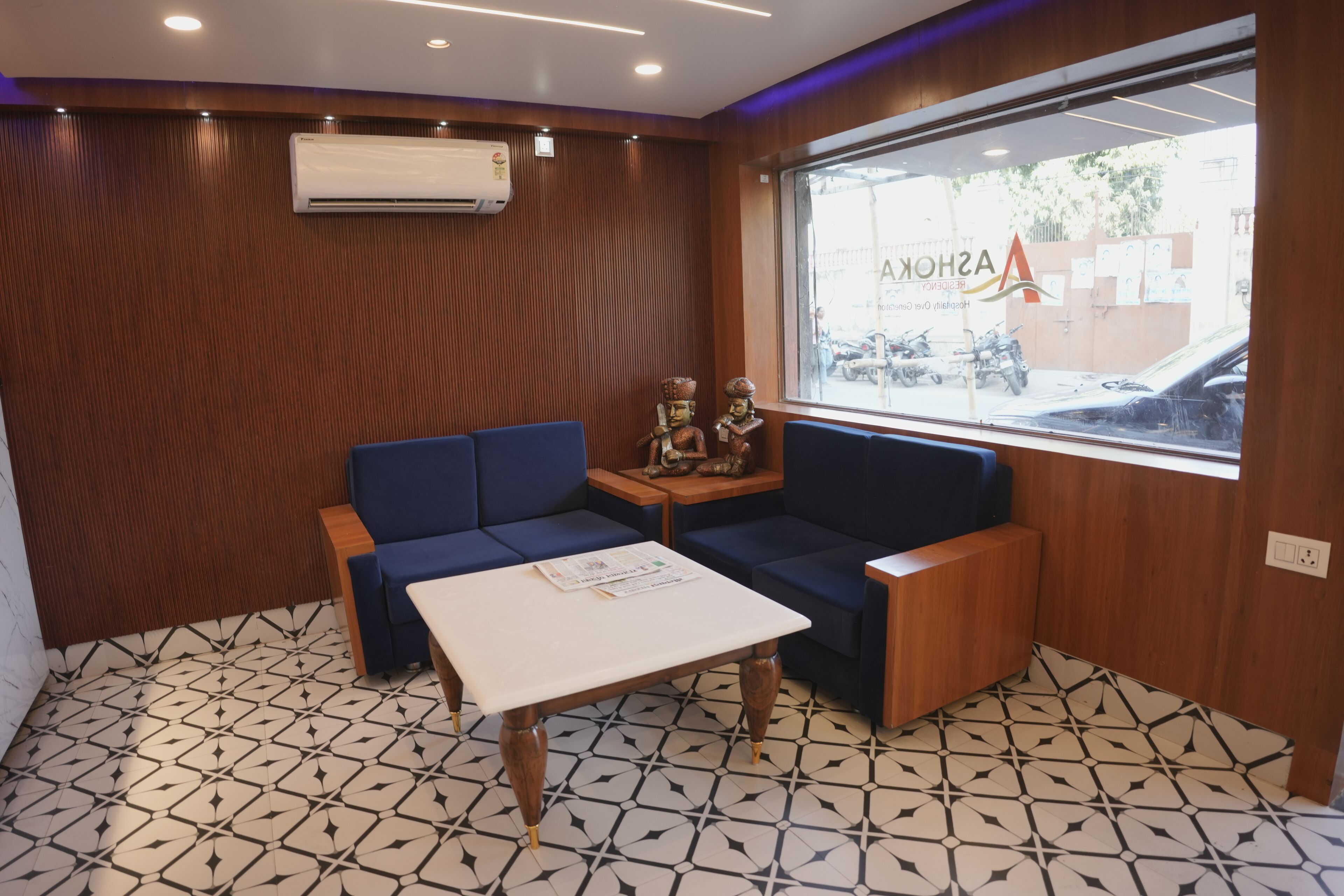 A room with a patterned floor, a blue sofa, a small table, and a window with a view of parked cars and a building outside.