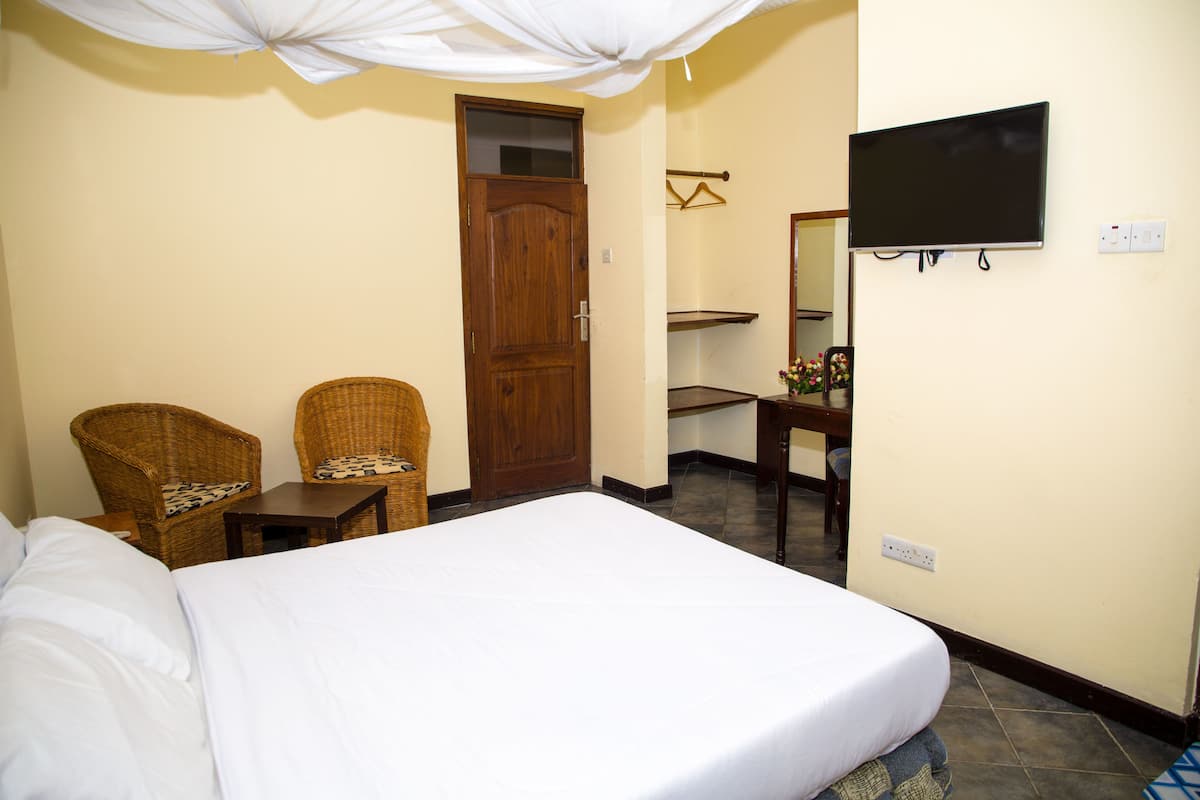 A hotel room with a bed, two wicker chairs, a wooden door, a television mounted on the wall, and a small table with a mirror and decorative items.
