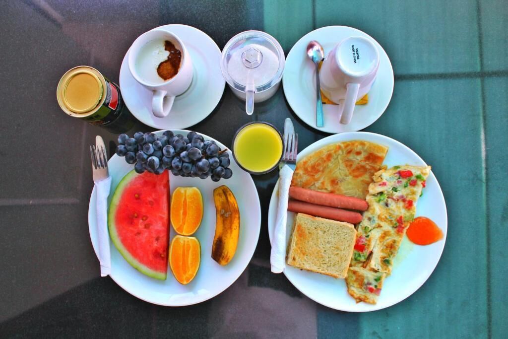 A breakfast setting with plates of food, including toast, hot dogs, and a slice of pizza, along with fruits like watermelon and bananas, and beverages.