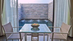 Et glasbord med to stole, en skål med frugt, og udsigt til et bassin gennem en stenvæg.