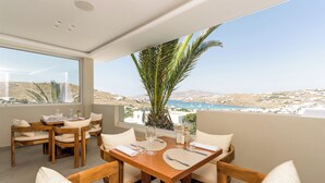 A dining area with a wooden table and chairs, a large window with a view of a coastal landscape. - Kallos of Mykonos (Mykonos)