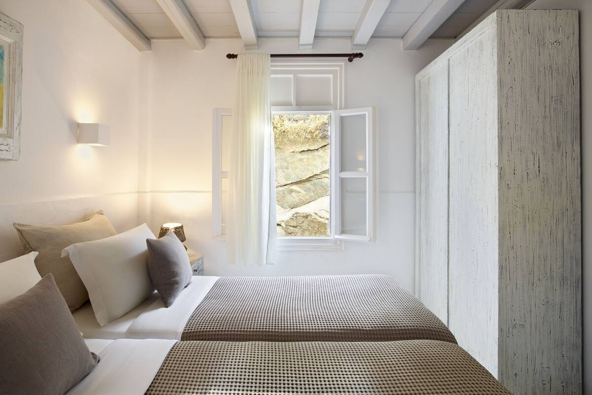 A bedroom with a bed, pillows, a bedside lamp, a window with a view of rocks, and a wooden wardrobe.