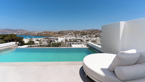 A modern pool area with a clear view of a coastal town and mountains in the background. - Kallos of Mykonos (Mykonos)