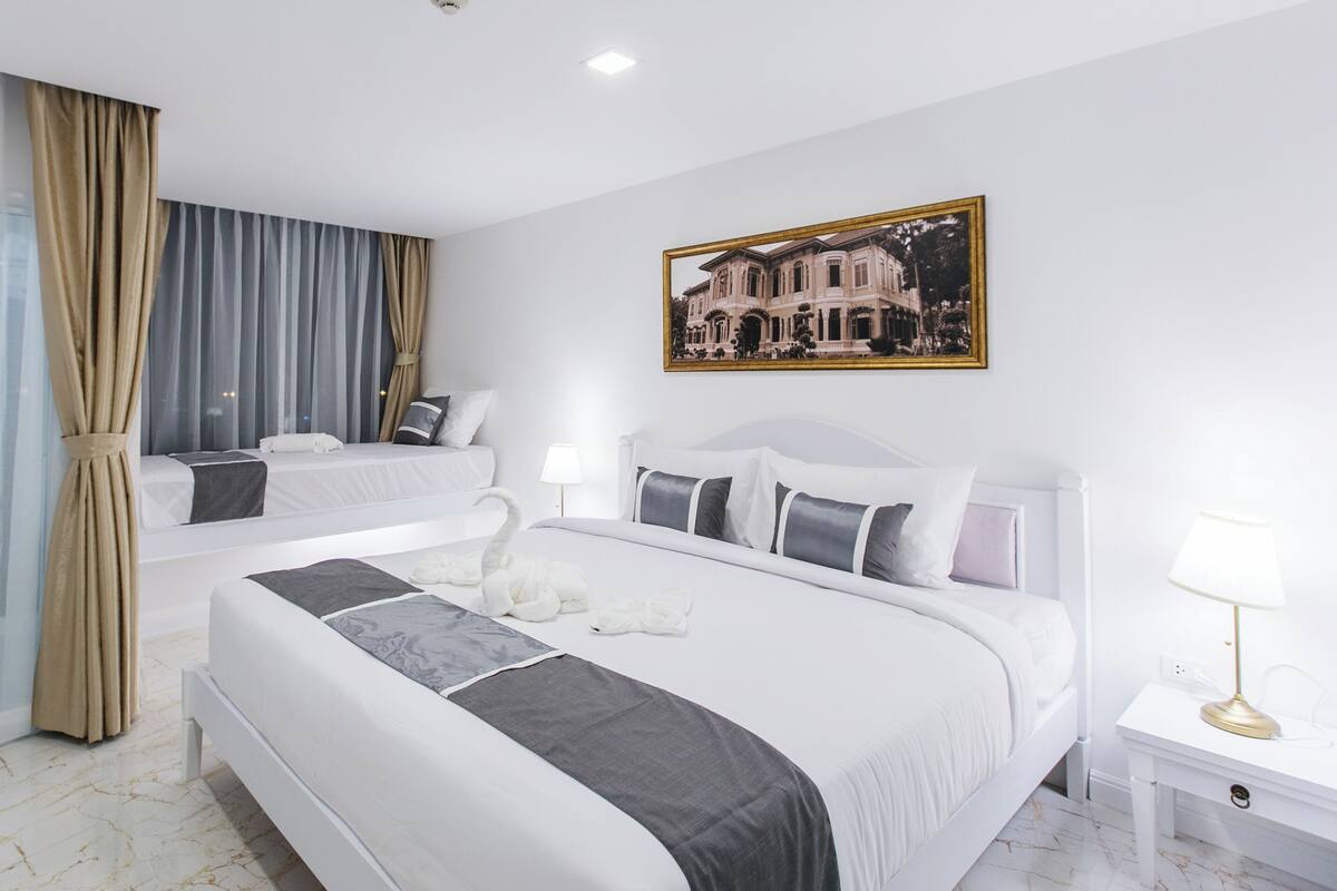 Habitación de hotel con dos camas, un ventanal con cortinas translúcidas, un cuadro de un edificio y una mesita de noche con lámpara.