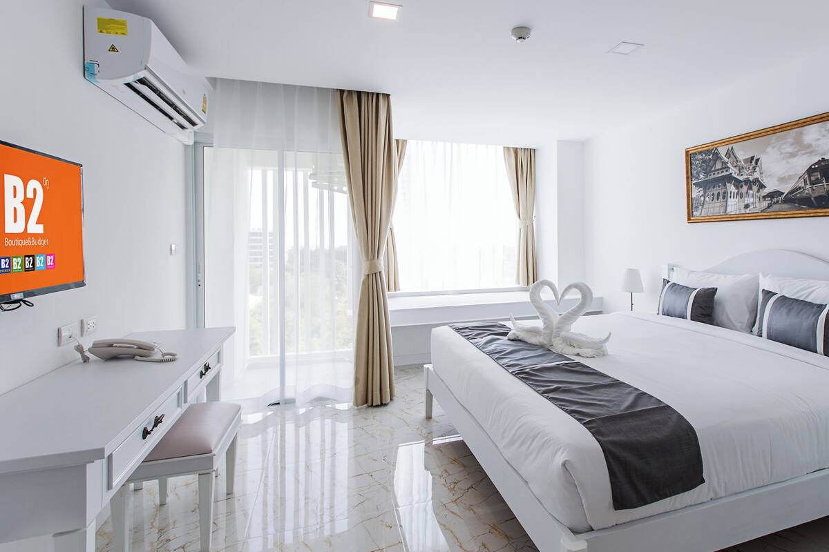 Una habitación de hotel moderna con una cama, un escritorio, una silla, un ventanal con cortinas y un cuadro en la pared.