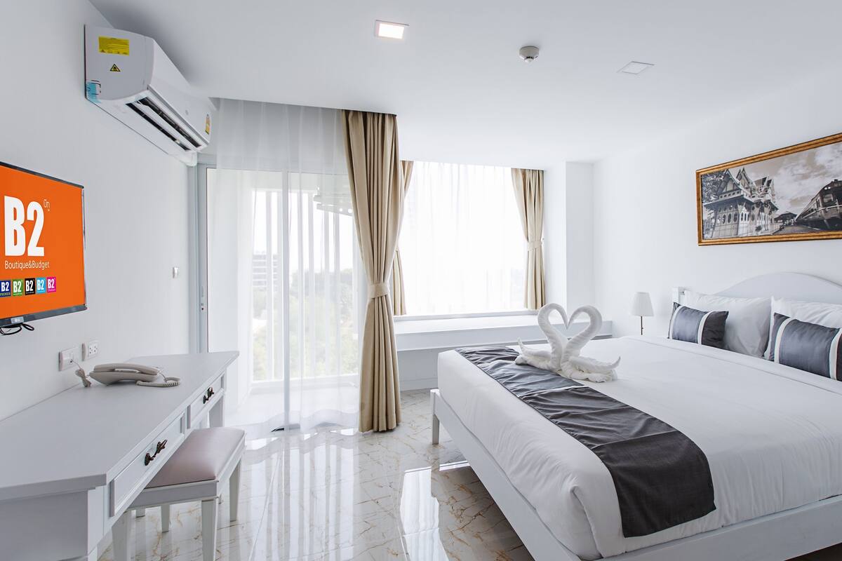 Una habitación de hotel con cama, escritorio, silla, teléfono, un cuadro y una ventana con cortinas.