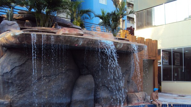 A swimming pool with a waterfall feature and people swimming. - Apartamento Grand Dolphin - Férias em Caldas Novas (Caldas Novas)