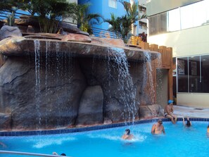 A swimming pool with a waterfall feature and people swimming. - Apartamento Grand Dolphin - Férias em Caldas Novas (Caldas Novas)