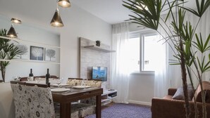 A dining area with a wooden table set for four, flanked by chairs with floral upholstery. A television is mounted on the wall, and a large window with sheer curtains is visible. - Apartamento Atalaia (Porto Alegre)