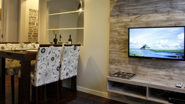 A modern living room with a flat-screen TV mounted on a textured wall, a wooden entertainment unit below, and a dining area with a table set for two. - Apartamento Atalaia (Porto Alegre)