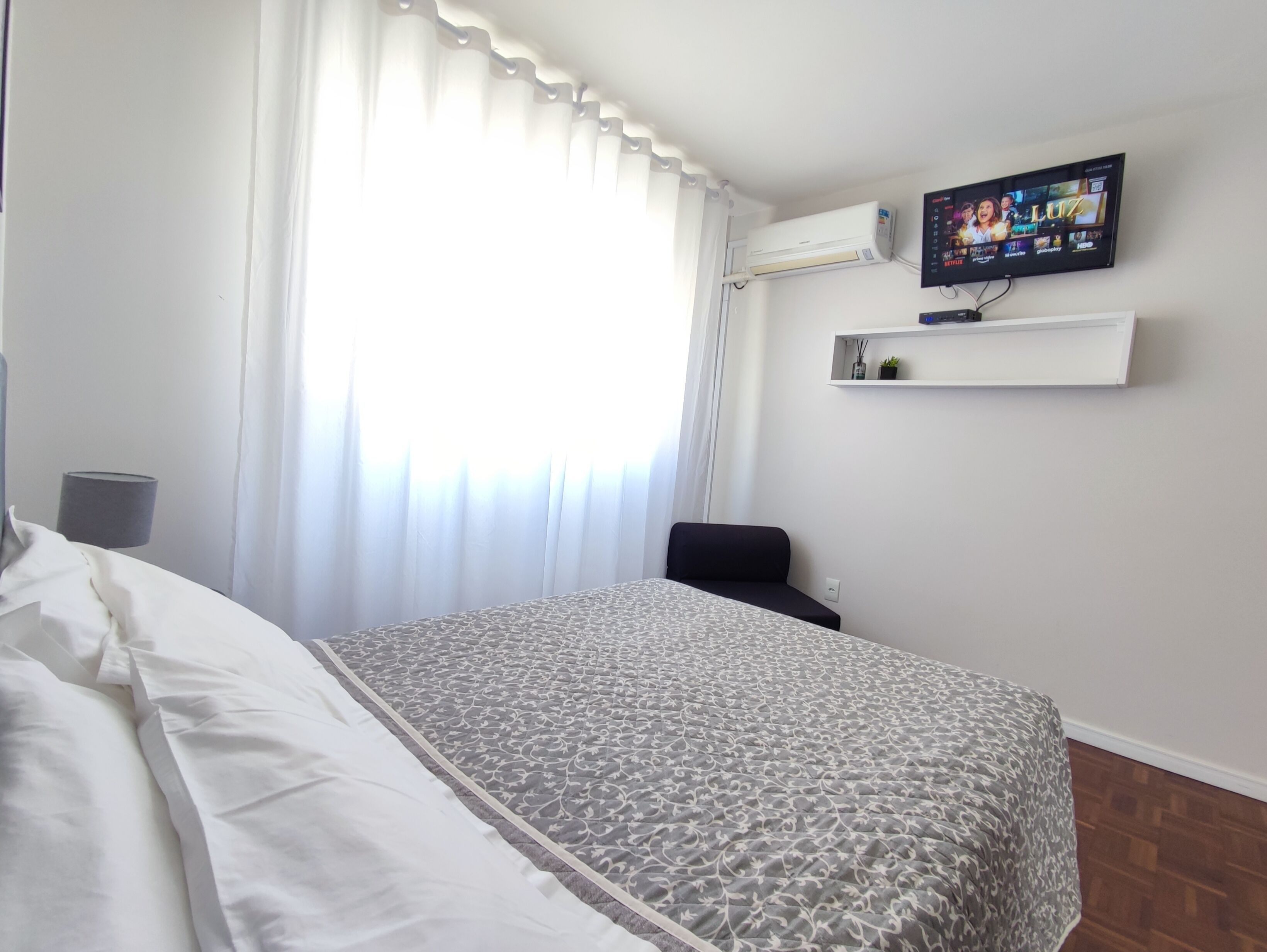 Quarto de hotel com uma cama, televisão na parede, aparelho de ar condicionado, prateleira pequena com objetos decorativos e janela com cortinas.