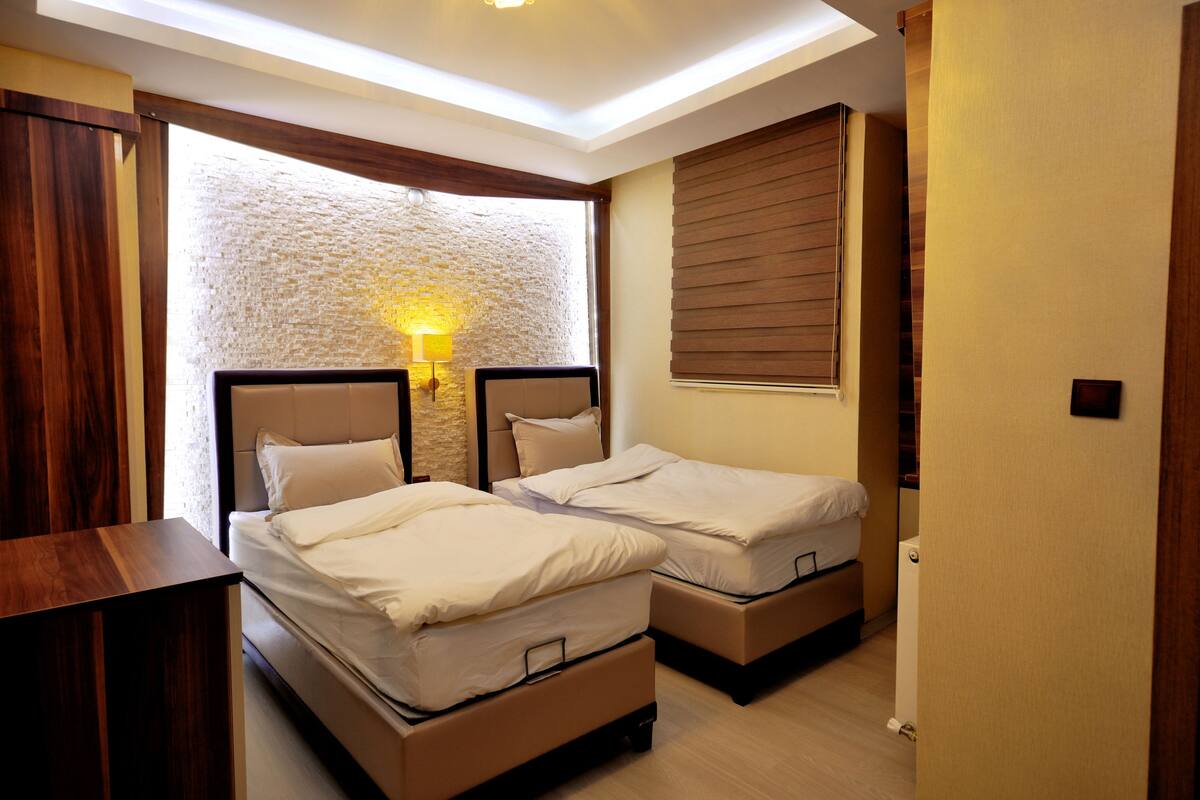 A bedroom with two beds, a wooden nightstand, a stone accent wall, and a window with blinds.