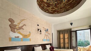 A hotel room with two beds, a large mural of ancient Egyptian figures, a circular ceiling design, and a balcony with a view. - El Nakhil Hotel (Luxor)