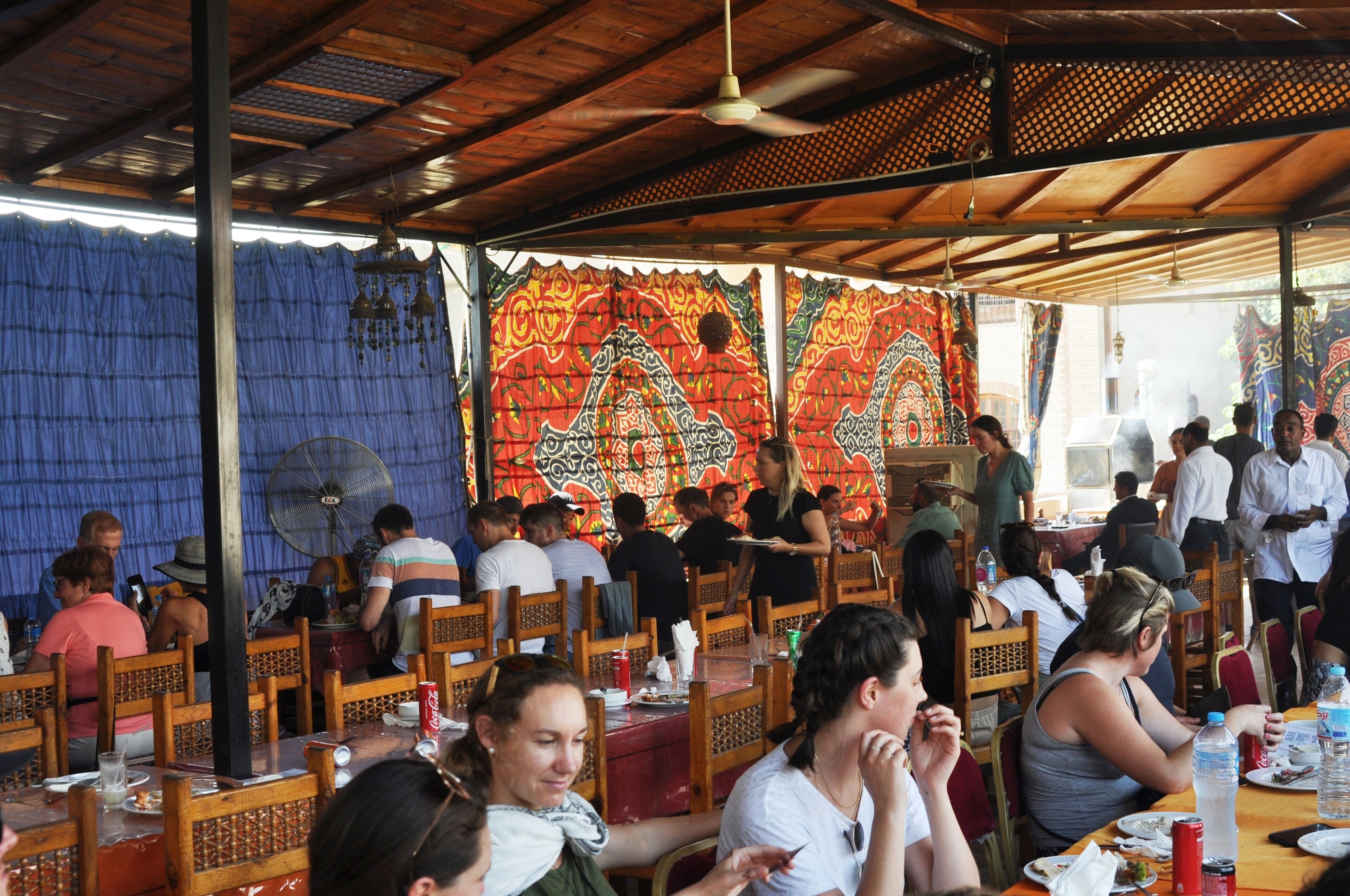 A crowded outdoor dining area with wooden tables and chairs, a ceiling fan, and patterned curtains.