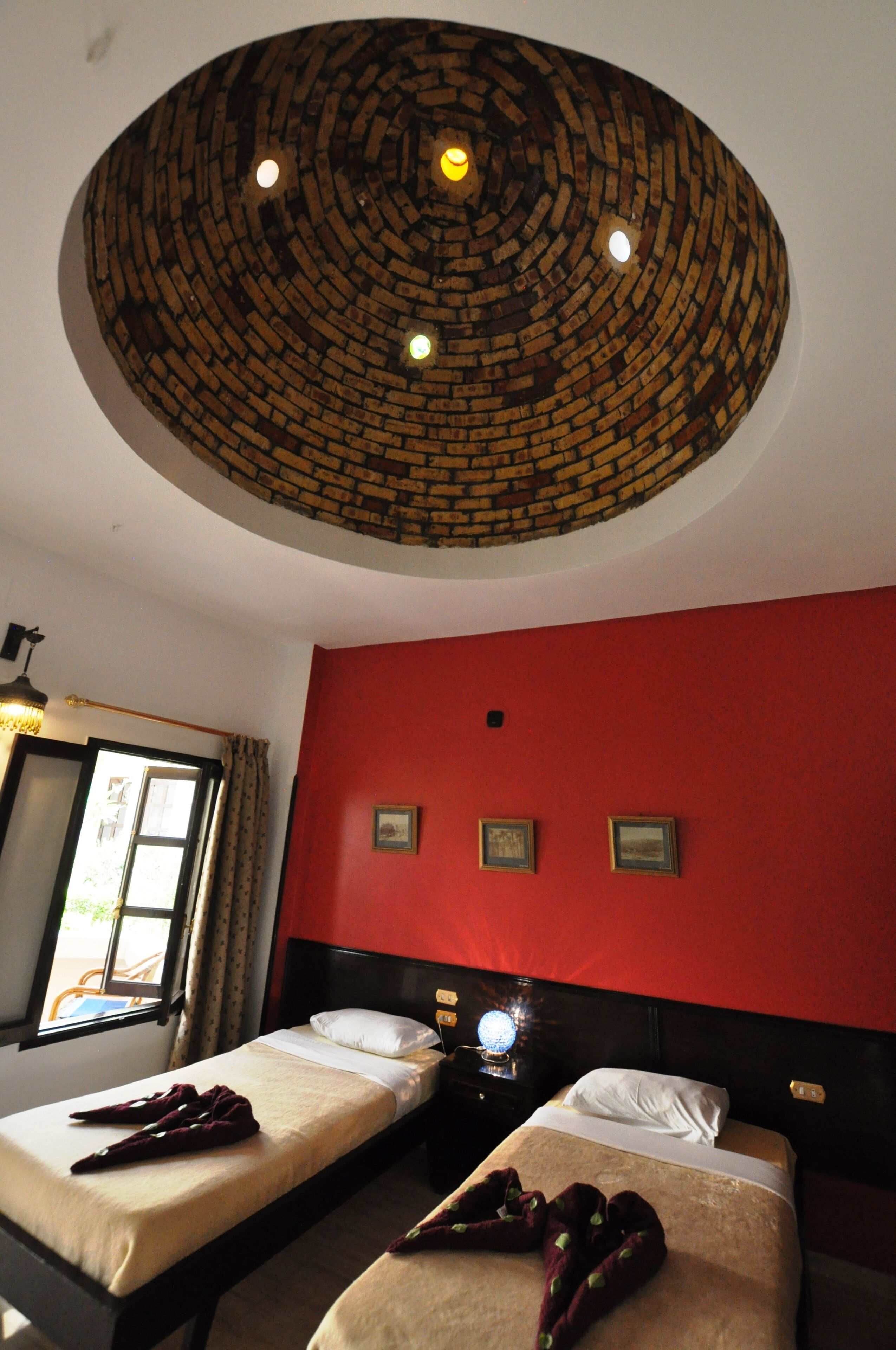 Une chambre d’hôtel avec deux lits, un mur d’accent rouge, un plafond à motifs de briques et une fenêtre avec des rideaux.