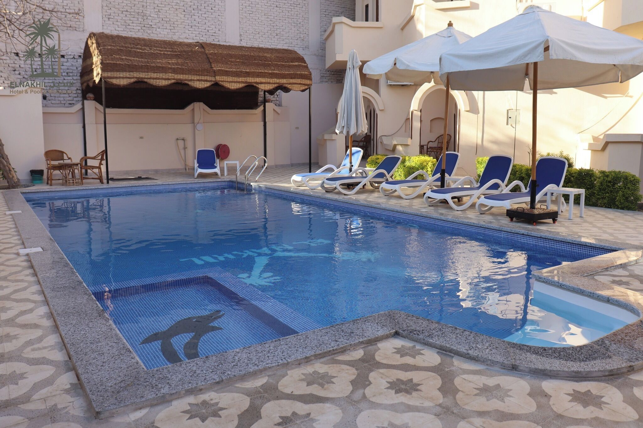 A swimming pool with lounge chairs and umbrellas, a tiled deck, and a building with a sign reading "EL NAKHIL Hotel & Pool".