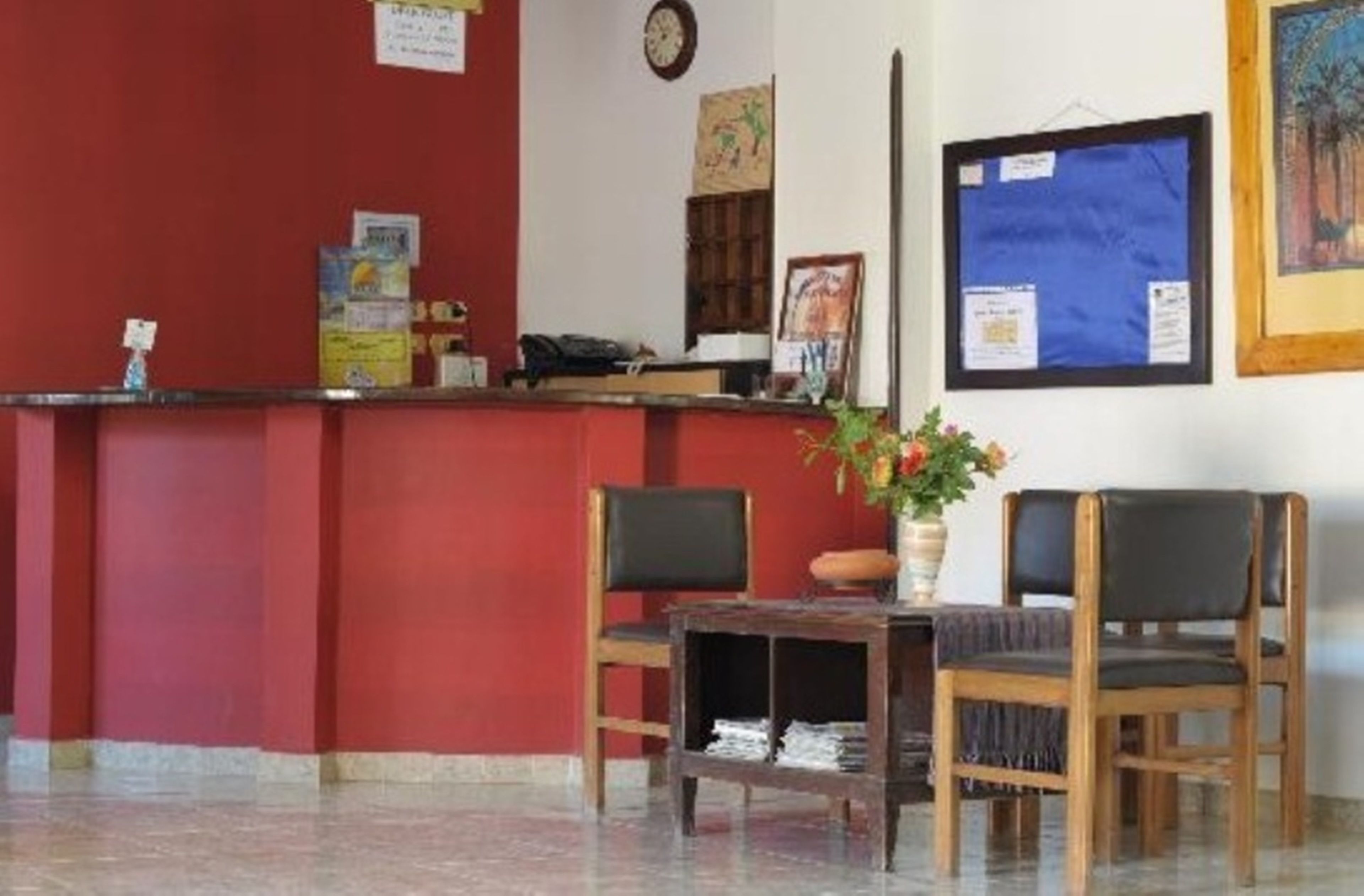 A reception area with a red wall, a reception desk, two chairs, a small table with a vase of flowers, and a clock on the wall.