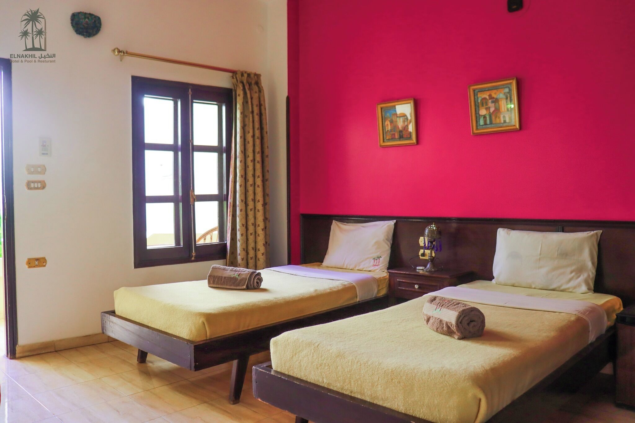 Habitación de hotel con dos camas individuales, una ventana con cortinas, una pared roja y dos cuadros enmarcados.