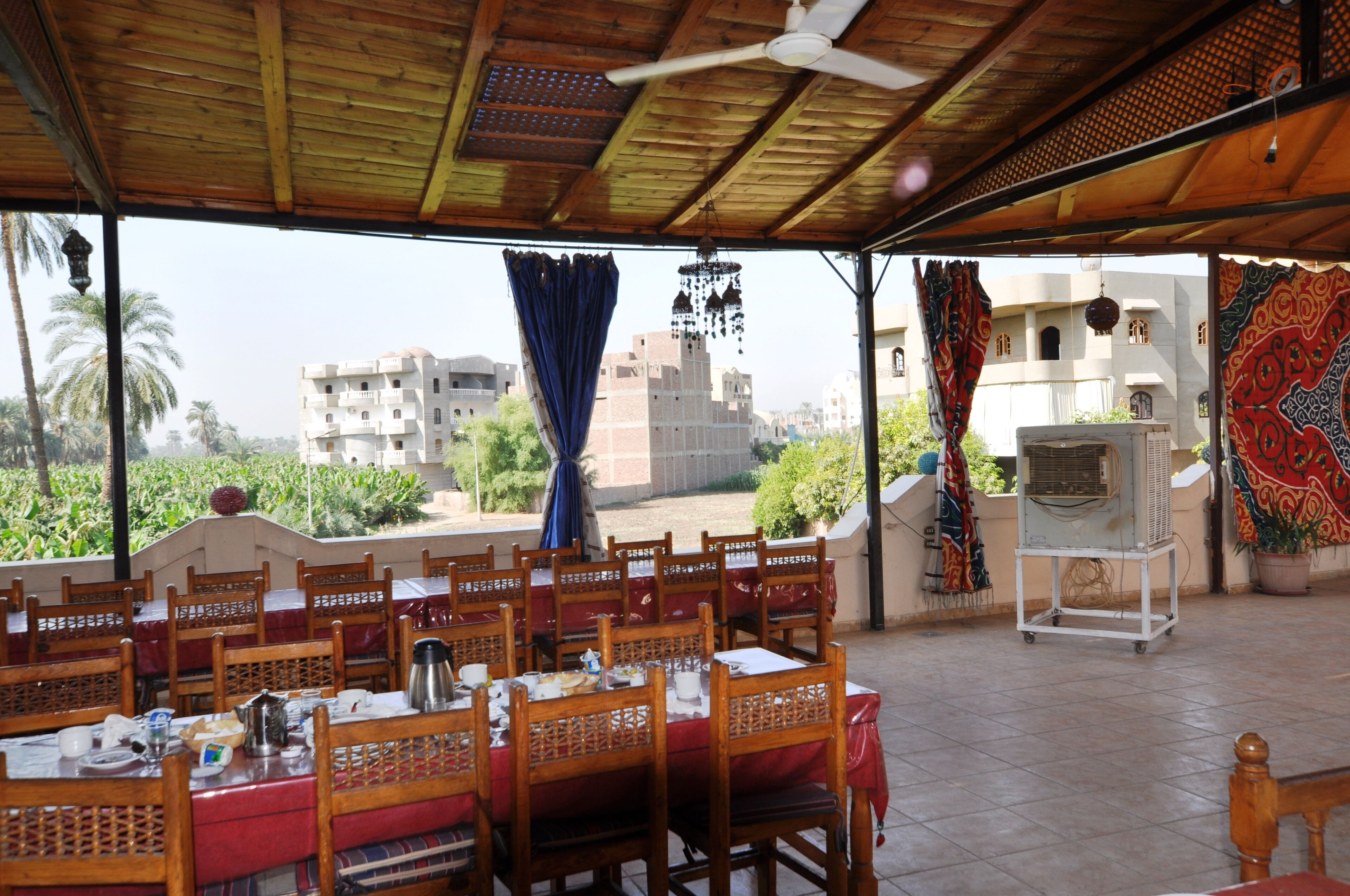 Un área de comedor al aire libre con mesas y sillas de madera, un ventilador de techo y vistas a edificios y follaje.