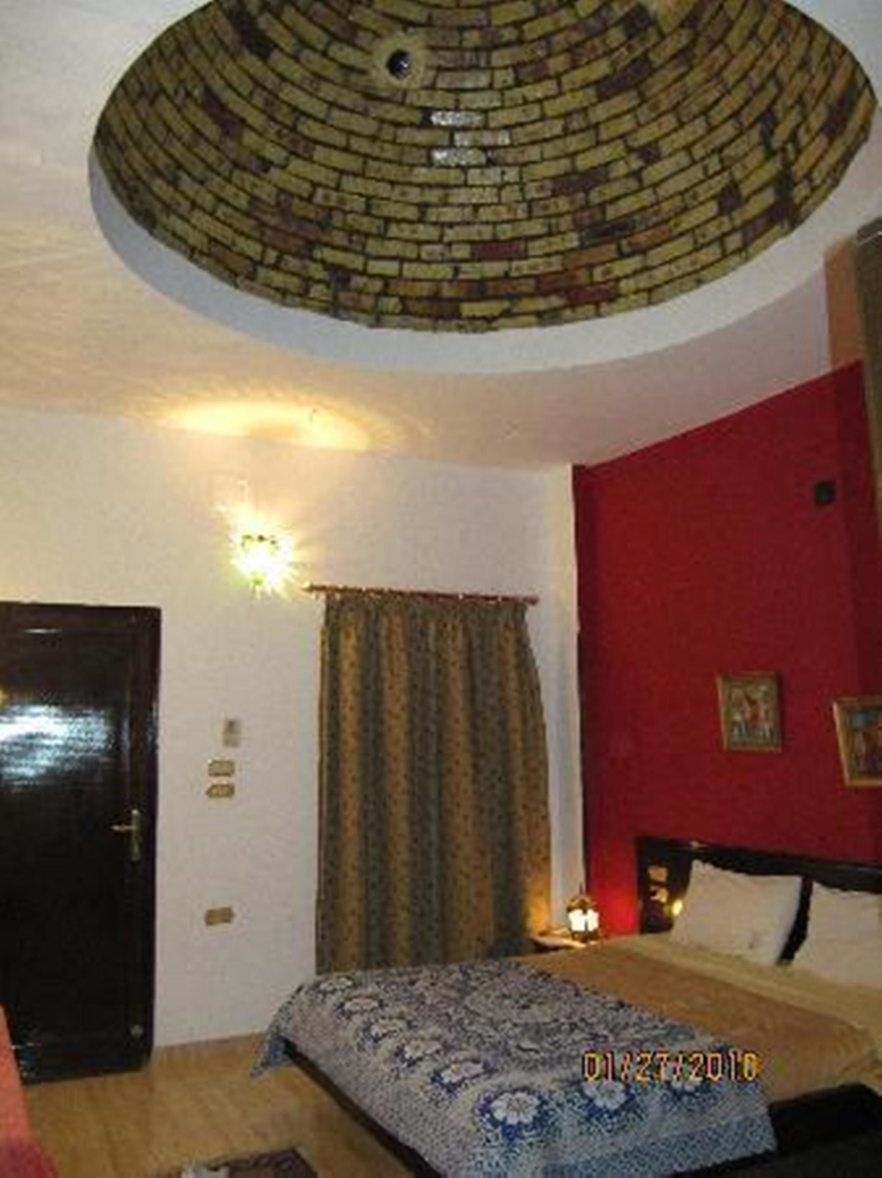 A room with a brick-patterned ceiling, a bed with a patterned bedspread, a red wall with a picture, and a door.