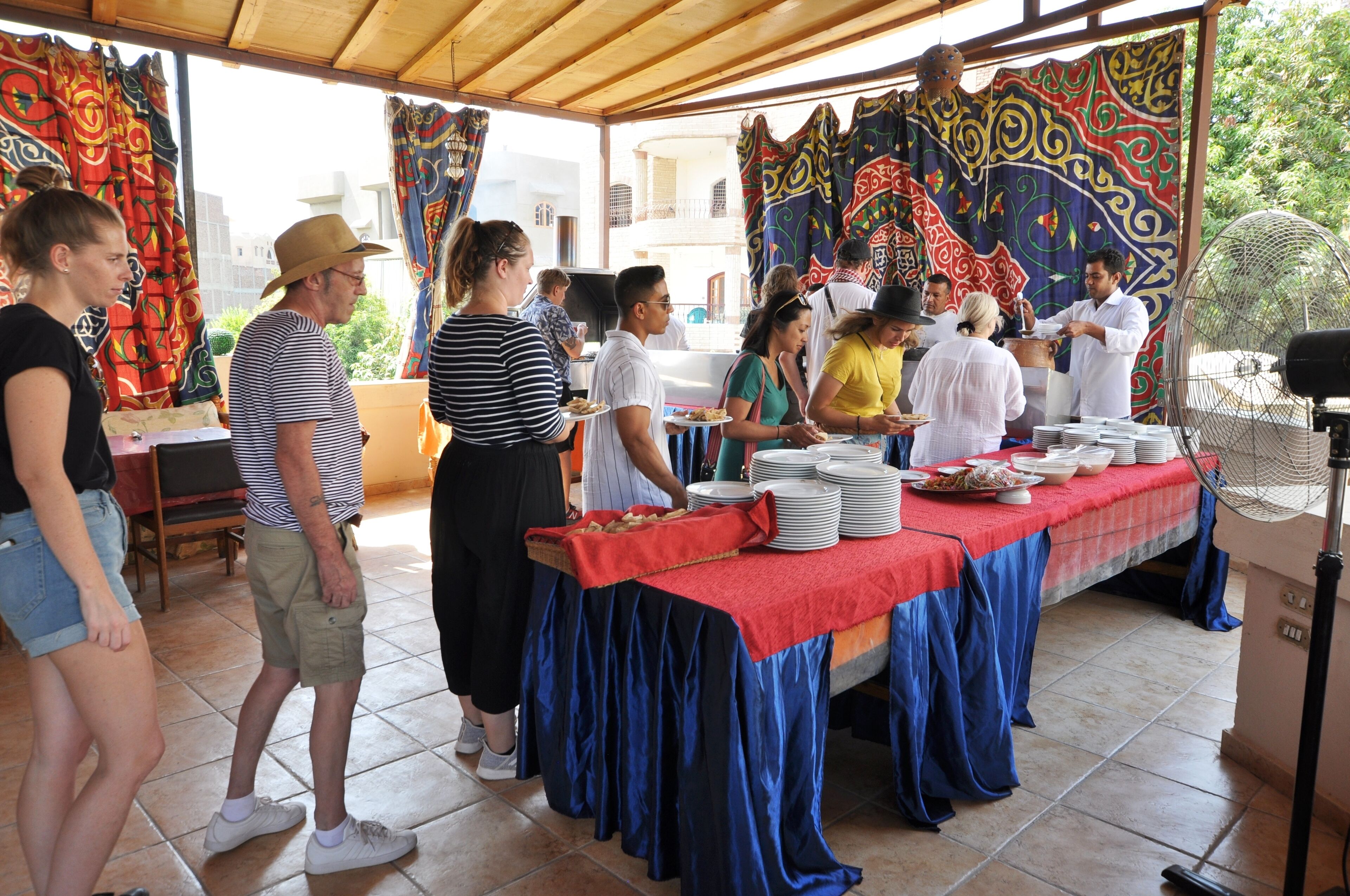 A covered outdoor dining area with a table set for a meal, surrounded by colorful curtains and a fan.