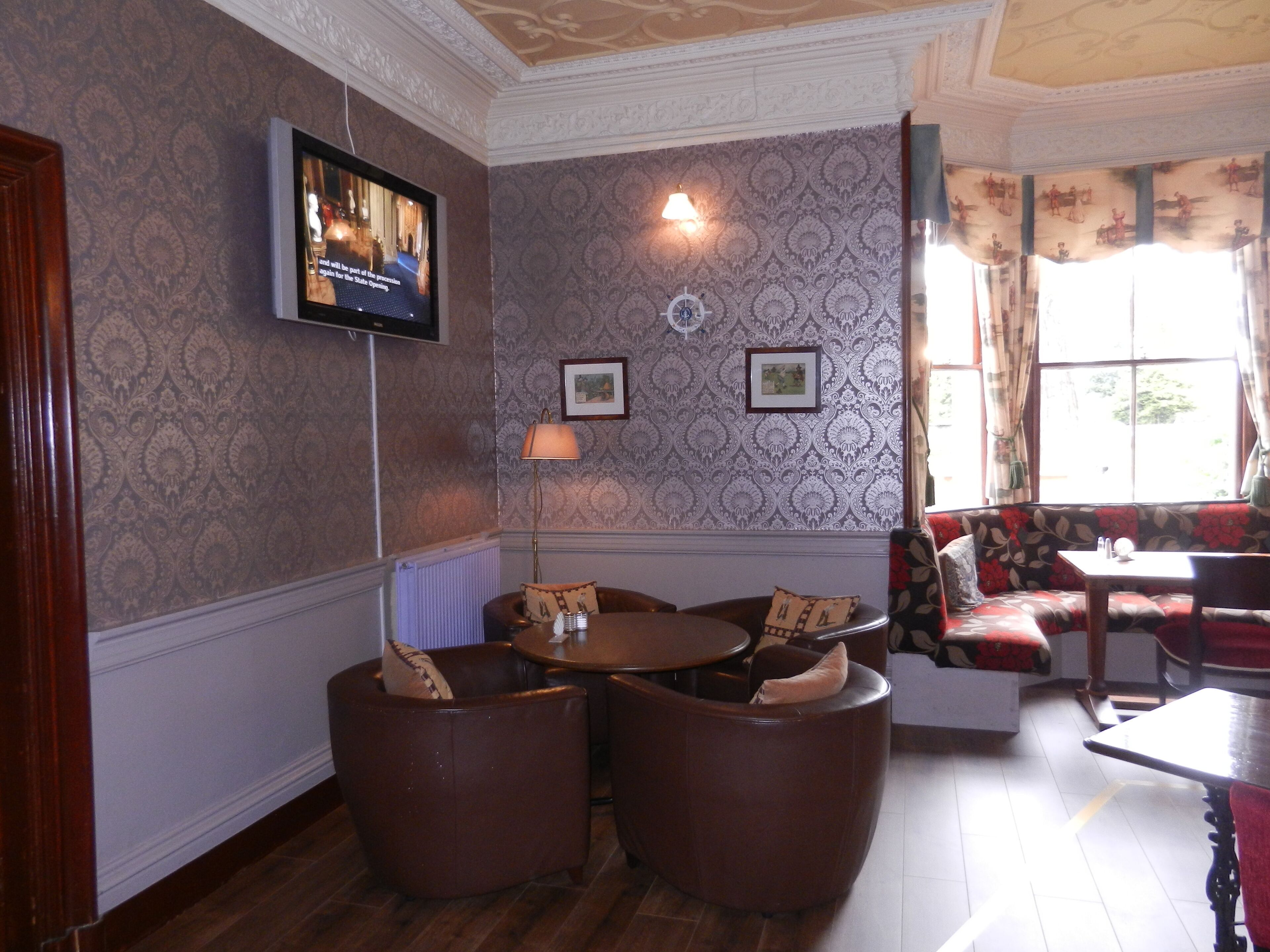 A room with patterned wallpaper, a television mounted on the wall, a floor lamp, and a seating area with a sofa and armchairs.