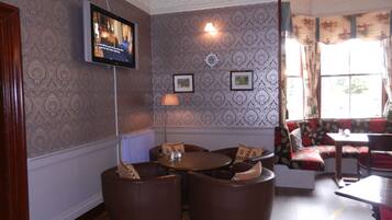 A room with patterned wallpaper, a television mounted on the wall, a floor lamp, and a seating area with a sofa and armchairs.