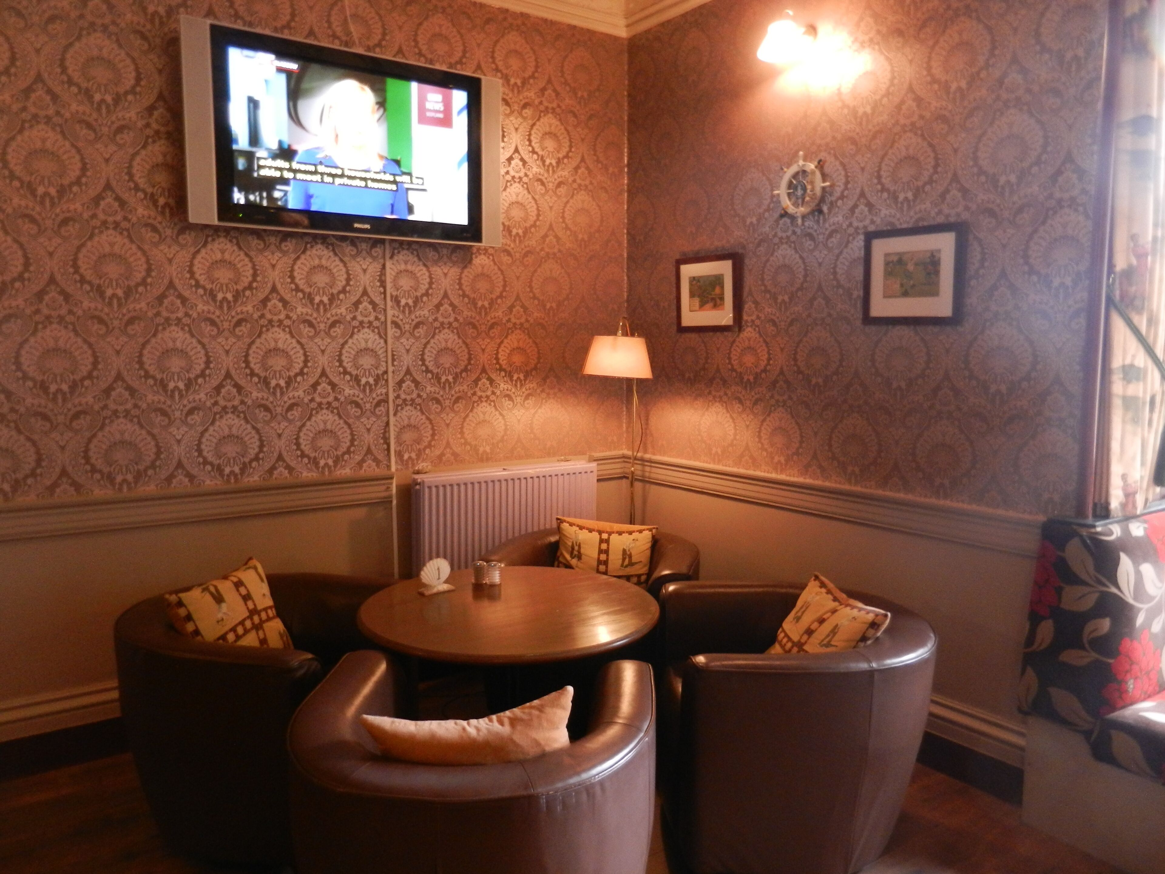 A corner room with a television mounted on the wall, patterned wallpaper, a radiator, and a small table with chairs.