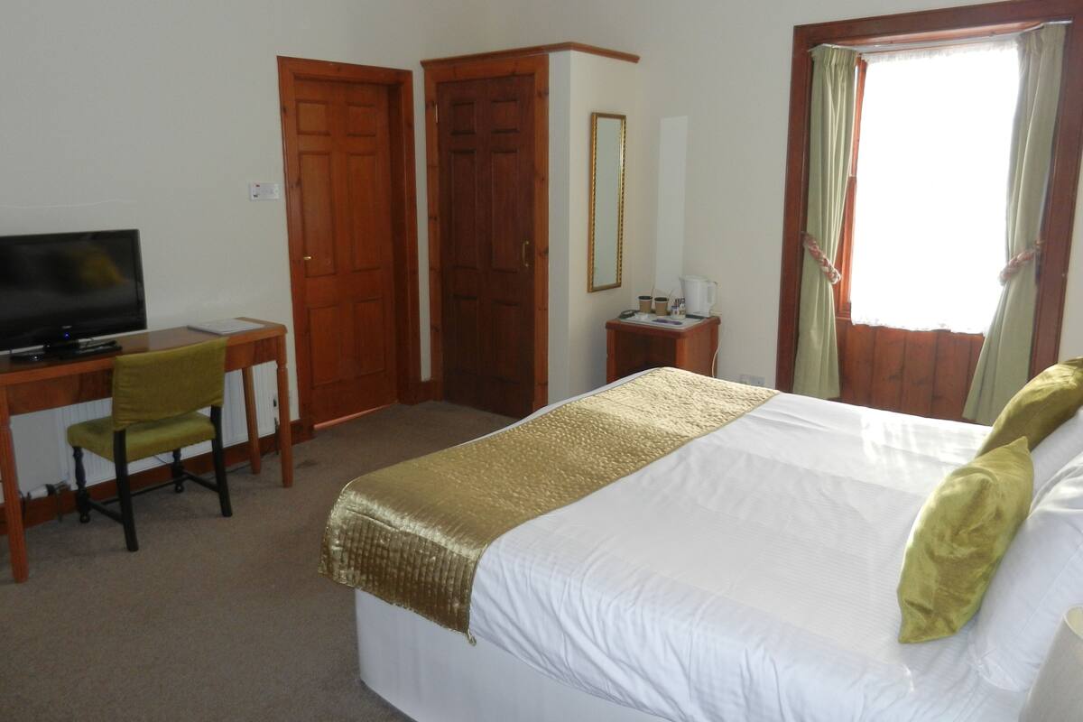 Habitación de hotel con cama, escritorio con silla, televisión, espejo y ventana con cortinas.