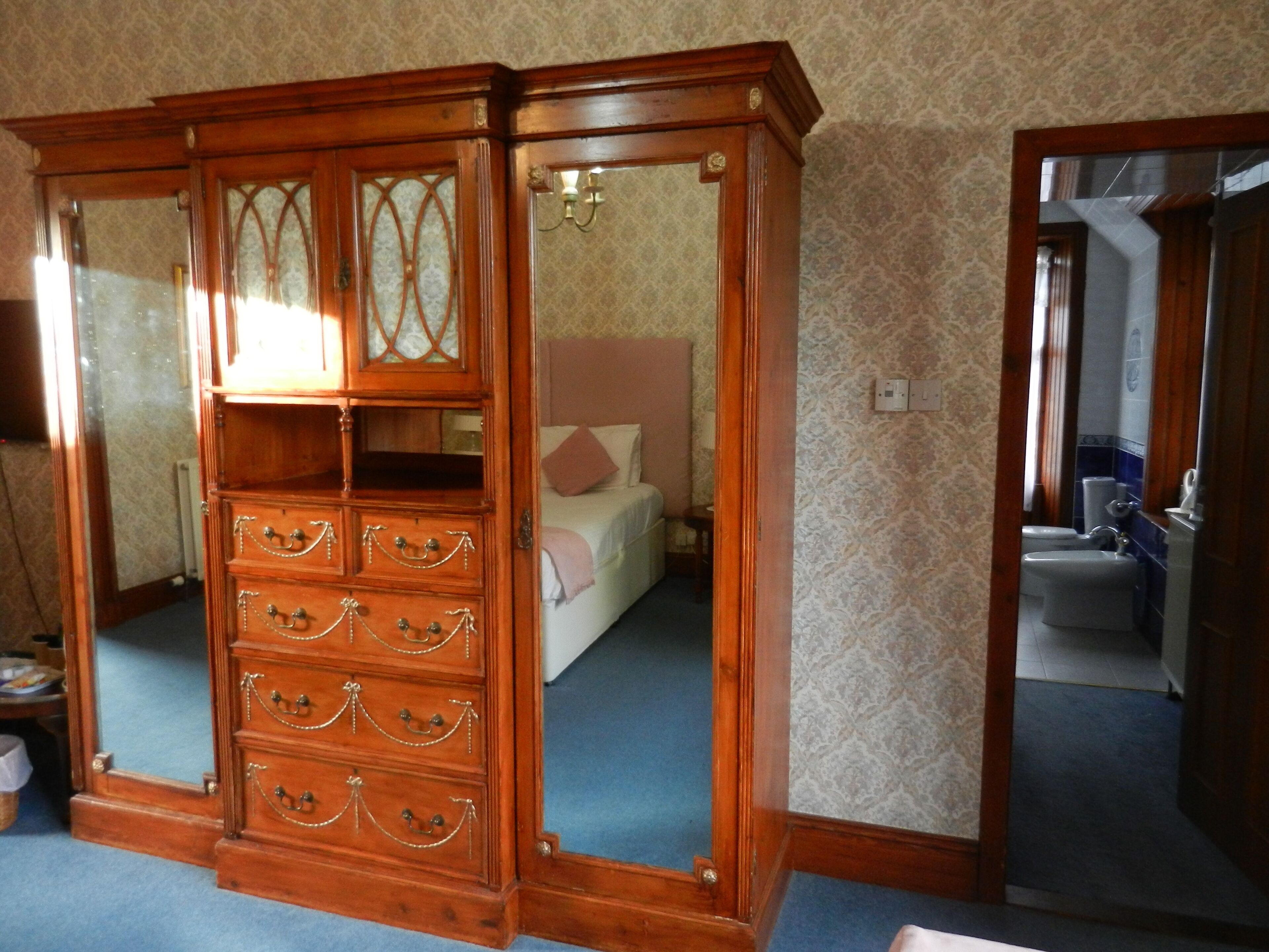 A wooden armoire with glass panels and multiple drawers, a bed with a pink pillow, and a bathroom visible through an open door.